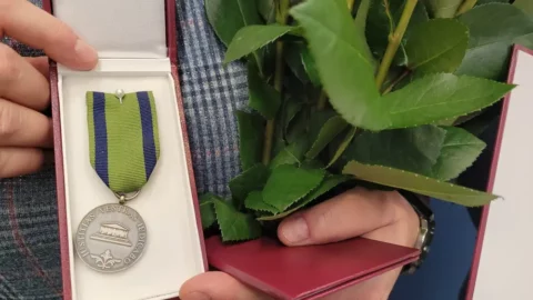 Osoba trzyma srebrny medal na zielonej wstążce, obok widać liście od róż.