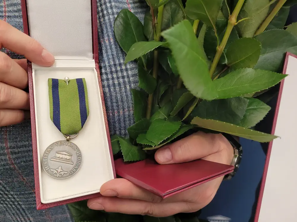 Osoba trzyma srebrny medal na zielonej wstążce, obok widać liście od róż.
